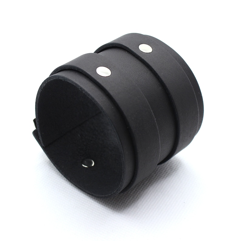 May include: A black leather cuff bracelet with silver metal accents. The bracelet is made of three layers of leather and has a simple, minimalist design.