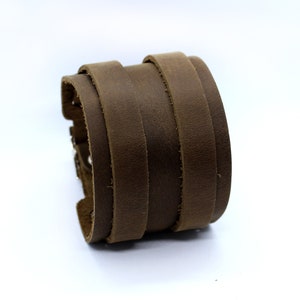 May include: A brown leather cuff bracelet with a layered design. The bracelet is made of multiple strips of leather that are stitched together.