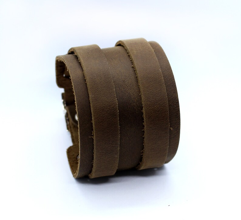 May include: A brown leather cuff bracelet with a layered design. The bracelet is made of multiple strips of leather that are stitched together.