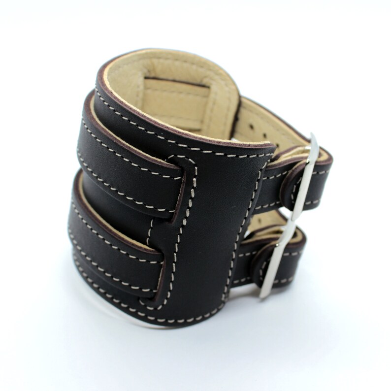 May include: A brown leather watch band with a silver buckle and white stitching. The band has two adjustable straps.