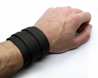 May include: A black leather cuff bracelet with a buckle closure. The bracelet is worn on a person's wrist.