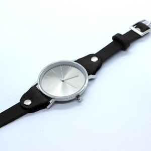Peut inclure: Une montre-bracelet argent&eacute;e avec un bracelet en cuir noir. Le cadran de la montre est argent&eacute; et pr&eacute;sente un design simple avec des aiguilles noires.