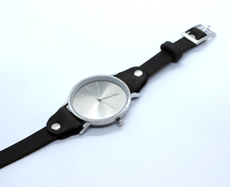 Peut inclure: Une montre-bracelet argent&eacute;e avec un bracelet en cuir noir. Le cadran de la montre est argent&eacute; et pr&eacute;sente un design simple avec des aiguilles noires.