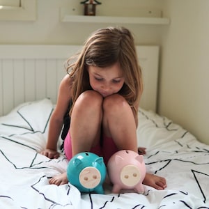 May include: A young girl sits on a bed with two piggy banks. One is light blue, the other is pink. Both have wooden snouts and small ears. The bed has a white comforter with a black geometric pattern.