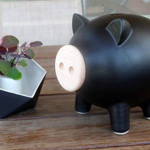 May include: A black piggy bank with a light wood-colored snout and small legs. Next to it is a geometric black and white planter with a small plant inside. The objects are on a wooden table.