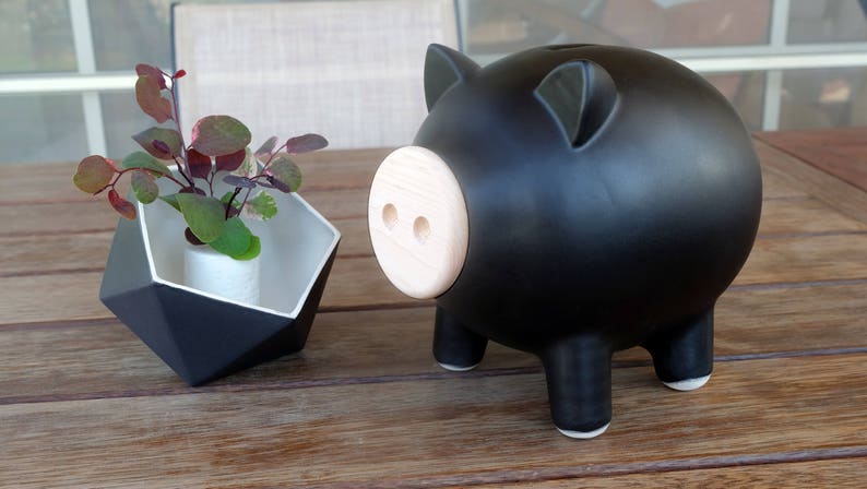 May include: A black piggy bank with a light wood-colored snout and small legs. Next to it is a geometric black and white planter with a small plant inside. The objects are on a wooden table.
