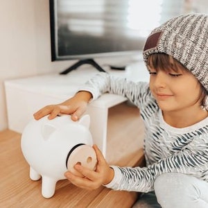 May include: A white ceramic piggy bank with a wooden nose. The piggy bank has four legs and two ears. The piggy bank is being held by a person wearing a striped shirt and a knit cap.