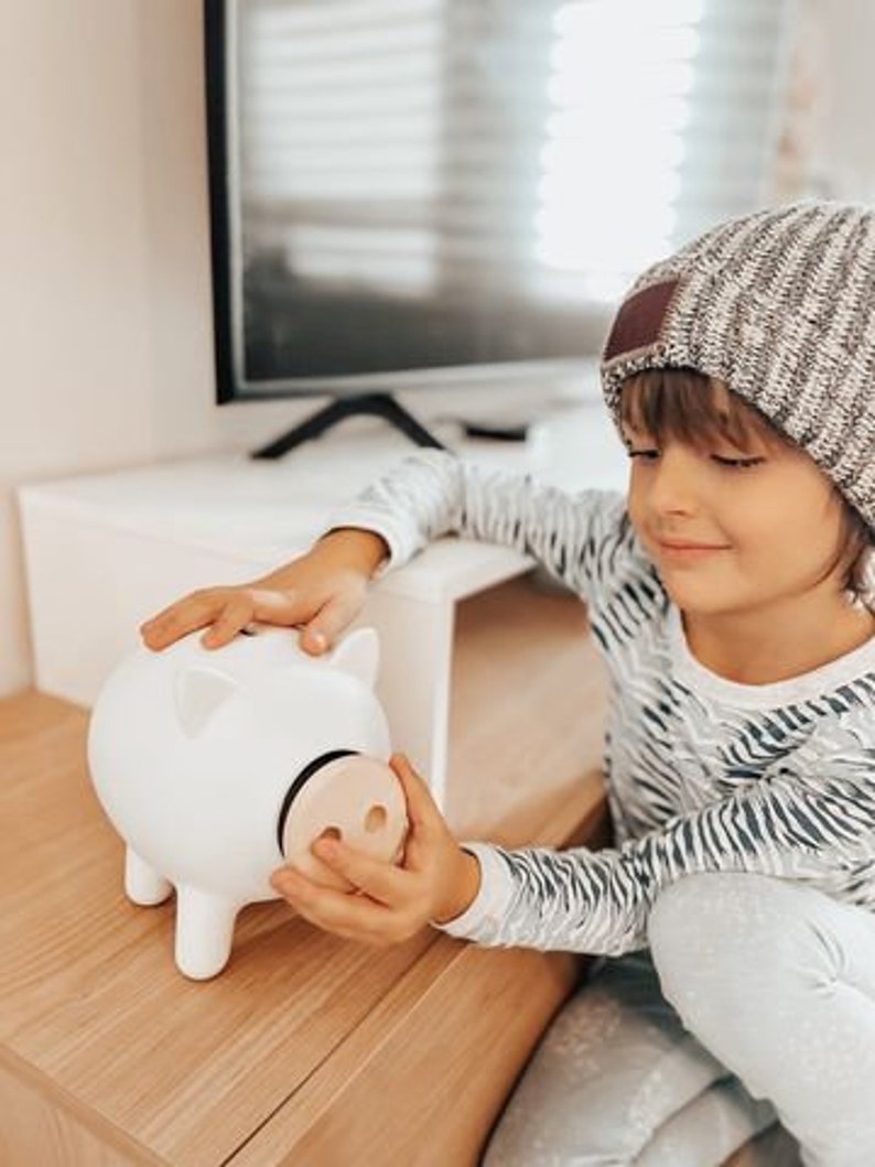 May include: A white ceramic piggy bank with a wooden nose. The piggy bank has four legs and two ears. The piggy bank is being held by a person wearing a striped shirt and a knit cap.