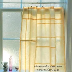 Pode incluir: Cortinas de janela transparentes amarelo claro com um padrão de grade de linhas amarelas mais escuras. As cortinas estão em frente a uma janela com vidro fosco. O texto "windowartcurtains.com norascurtains.com" é visível.