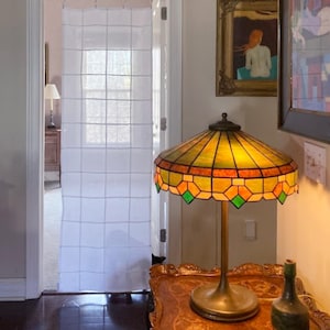 May include: A white sheer curtain with a grid pattern hangs in a doorway. In the foreground, a stained glass lamp with a yellow and green geometric shade sits on a wooden table next to a small green vase, with framed art on the wall.