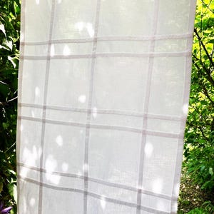 May include: A semi-sheer white curtain panel featuring a subtle grid pattern formed by thin, pale pink vertical and horizontal lines. The fabric has a visible woven texture, allowing light to pass through the material in a soft, diffused manner.