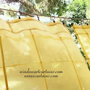 Pode incluir: Cortinas amarelas transparentes com um padrão quadriculado, penduradas em anéis pretos em uma corda. As cortinas são ligeiramente translúcidas, permitindo a passagem da luz. O texto "windowartcurtains.com norascurtains.com" é visível na parte inferior.