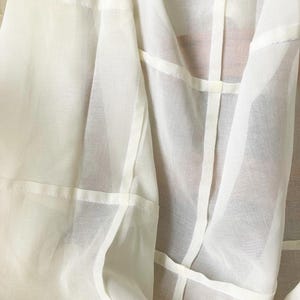 May include: A sheer off-white garment featuring a lightweight, translucent fabric construction. The piece is detailed with visible vertical and horizontal seams that create a structured grid pattern across the surface of the material.