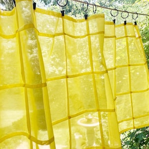 May include: A bright yellow sheer curtain panel featuring a grid pattern of horizontal and vertical fabric strips. The curtain is suspended from a thin wire by several black metal rings with clips, allowing the light to pass through the fabric.