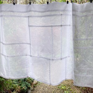 May include: A white sheer curtain panel featuring a geometric grid pattern created by opaque fabric strips. The lightweight, semi-transparent textile displays a series of rectangular and square sections arranged in a structured, linear composition.