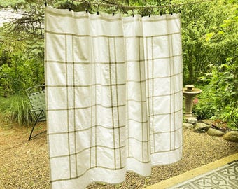 May include: A white shower curtain featuring a grid pattern of thin, muted olive green lines. The curtain is suspended from a horizontal wire by a series of dark metal rings, creating vertical folds across the fabric surface.
