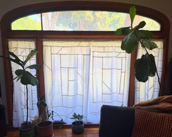 May include: A set of white sheer curtains featuring a geometric grid pattern of thin dark lines, hung on a rod within a large arched wooden window frame, accompanied by several potted fiddle leaf fig plants placed on the floor in front of the glass.