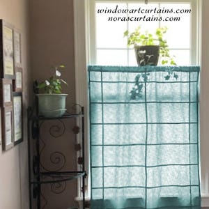 以下が含まれることがあります： 窓の前に、格子模様のターコイズ色の窓カーテンが吊るされています。カーテンはシアー生地で作られています。「windowartcurtains.com norascurtains.com」というテキストが窓に見えます。鉢植えの植物が窓台に置かれています。