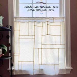 Pode incluir: Uma cortina de janela branca com um padrão de grade geométrica em bege. A cortina está pendurada em frente a uma janela. O texto "windowartcurtains.com norascurtains.com" é visível na janela.