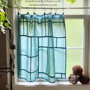 Puede incluir: Una cortina transparente azul claro con un diseño de cuadrícula cuelga en una ventana. La cortina está suspendida de una barra con anillos. El texto "windowartcurtains.com norascurtains.com" es visible. Esferas decorativas en el alféizar.