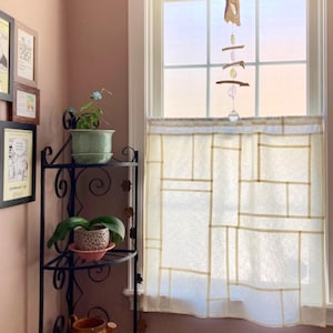 May include: A white cafe curtain featuring a geometric grid pattern of thin, light tan lines. The curtain is hung on a window, with a hanging mobile made of driftwood, crystals, and a clear glass sphere suspended in front of the window pane.