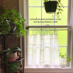 May include: A white cafe curtain with a geometric grid pattern hangs from a white tension rod with circular rings. The text windowartcurtains.com and norascurtains.com is visible on the window glass above the curtain in black font.