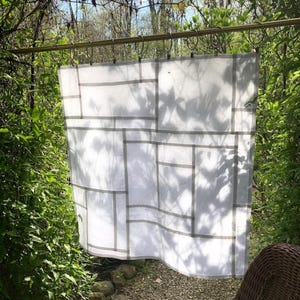 May include: A white fabric curtain featuring a geometric pattern of thin, intersecting gray lines forming various rectangular shapes. The curtain is suspended from a horizontal rod by a series of metal rings with clips along the top edge.
