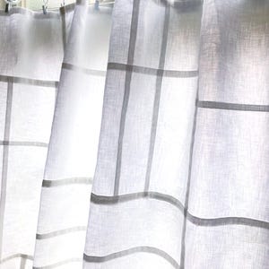 May include: A white semi-sheer curtain panel featuring a subtle grid pattern of horizontal and vertical lines. The fabric is suspended from a white rod by a series of white circular rings with small metal clips attached to the top edge of the fabric.