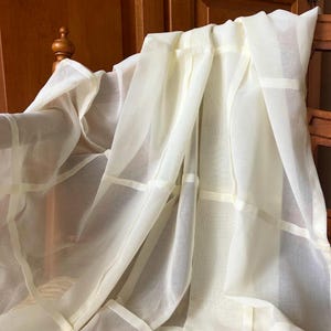 Pode incluir: Painel de cortina transparente creme com um padrão de grade. A cortina é drapeada sobre uma estrutura de madeira, com as dobras do tecido criando sombras e textura. A cortina é feita de um material leve e semitransparente.