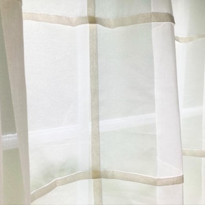 May include: A sheer white curtain panel featuring a grid pattern created by opaque, light beige horizontal and vertical fabric bands. The lightweight, translucent material allows soft light to filter through, highlighting the geometric design.