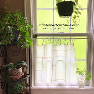 May include: A white cafe curtain with a geometric grid pattern hangs from a white tension rod with circular rings. The text windowartcurtains.com and norascurtains.com is visible on the window glass above the curtain in black font.