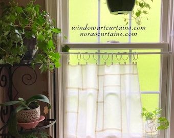 May include: A white cafe curtain with a geometric grid pattern hangs from a white tension rod with circular rings. The text windowartcurtains.com and norascurtains.com is visible on the window glass above the curtain in black font.