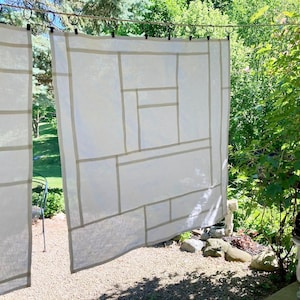 May include: Two off-white, semi-sheer curtains featuring a grid-like pattern of thin, light beige horizontal and vertical lines. The curtains are suspended from a thin metal rod by small black clips and circular rings, creating a geometric design.