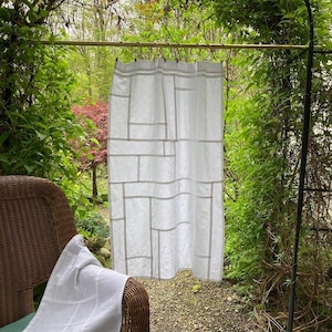 May include: White curtain with a geometric design hanging outdoors. The curtain has a grid pattern of light gray lines. A white cloth is draped over a wicker chair. The background features lush greenery and an arched structure.