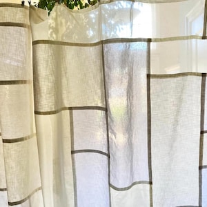 May include: A semi-sheer curtain panel featuring a grid-like pattern of intersecting horizontal and vertical lines in a muted taupe color against an off-white fabric base. The fabric has a visible weave texture and hangs in soft, vertical folds.