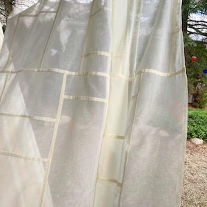 Pode incluir: Cortinas transparentes creme com um padrão de grade de fitas bege claro. As cortinas estão penduradas ao ar livre, com vista para um gramado verde e um corpo de água ao fundo. O tecido parece leve e arejado.