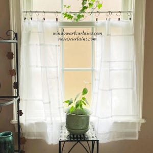 Pode incluir: Cortinas brancas transparentes com um padrão de grade penduradas em frente a uma janela. O texto "windowartcurtains.com norascurtains.com" é visível nas cortinas. Uma pequena planta verde em vaso está em um suporte de metal preto em frente à janela.
