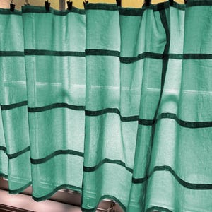 May include: A light green semi-sheer curtain panel featuring horizontal dark green stripes. The curtain is suspended from a white rod by a series of black metal rings with clips, allowing the fabric to hang in soft, vertical folds across the window.