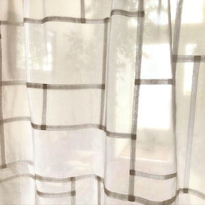 May include: A sheer white curtain featuring a grid pattern of thin, light gray horizontal and vertical lines. The fabric is translucent, allowing light to pass through, and the geometric design creates a series of rectangular shapes across the panel.