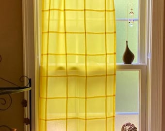 May include: A bright yellow window curtain featuring a grid pattern of darker yellow lines. The curtain is suspended from a white tension rod using silver metal rings with clips, covering a portion of a window pane with a light-filtering fabric.