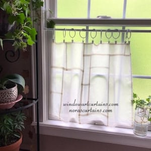 Pode incluir: Cortinas de janela brancas transparentes com um padrão quadriculado em marrom claro. As cortinas penduram-se de argolas de metal numa barra. O texto "windowartcurtains.com norascurtains.com" está impresso nas cortinas. As plantas são visíveis.