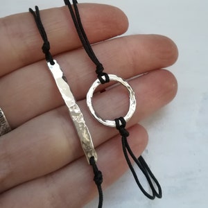 Puede incluir: Dos pulseras minimalistas con cordón negro y dijes de plata. Un dije es una barra fina martillada y el otro es un círculo.