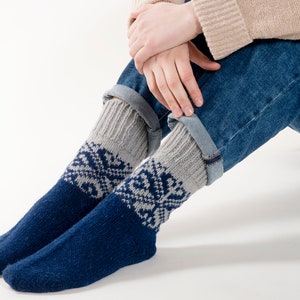 Könnte beinhalten: Ein Paar blau-graue Stricksocken mit Schneeflockenmuster. Die Socken werden mit hochgekrempelten Jeans getragen. Die Socken haben einen dunkelblauen Fuß und eine graue Manschette mit weißem Schneeflocken-Design.