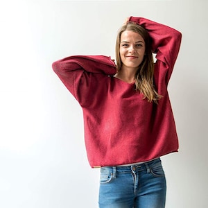 May include: A woman wearing a red sweater with long sleeves and blue jeans. She is standing in front of a white wall.