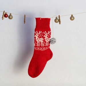 Puede incluir: Un calcetín navideño rojo tejido con un patrón de renos blancos cuelga de una cuerda con pequeños adornos. El calcetín tiene un pompón y se muestra sobre un fondo blanco.