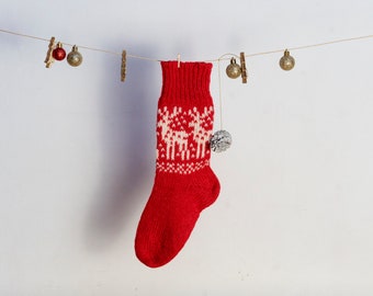 Puede incluir: Un calcetín navideño rojo tejido con un patrón de renos blancos cuelga de una cuerda con pequeños adornos. El calcetín tiene un pompón y se muestra sobre un fondo blanco.