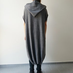 May include: A gray hooded tunic dress with a unique, rounded hemline. The sleeveless dress is made of a soft, knitted material. The model is wearing black pants and black boots.