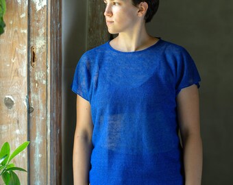 May include: A person wearing a blue, sheer, short-sleeved shirt and black shorts. They are standing near a window with wooden trim.