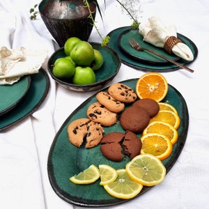 May include: A teal ceramic serving platter with an oval shape, holding chocolate chip cookies, chocolate cookies, orange slices, and lemon slices. Green apples are in a teal bowl. Teal plates and a vase are also present.