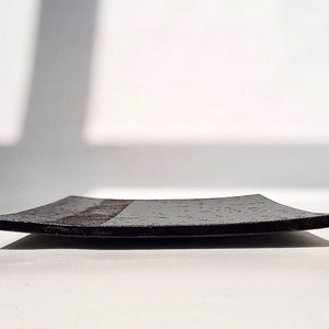 May include: A black square ceramic serving tray with a textured surface. The tray has a slightly curved edge.